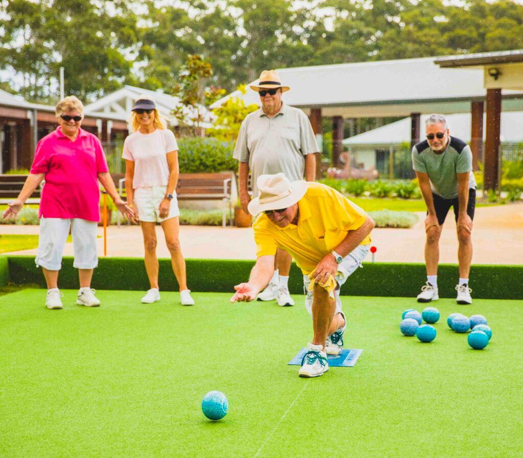 Man in yellow shirt rolls a blue bowling ball on a green lawn in a lifestyle village, while four others watch a casual game in a sunny outdoor setting.