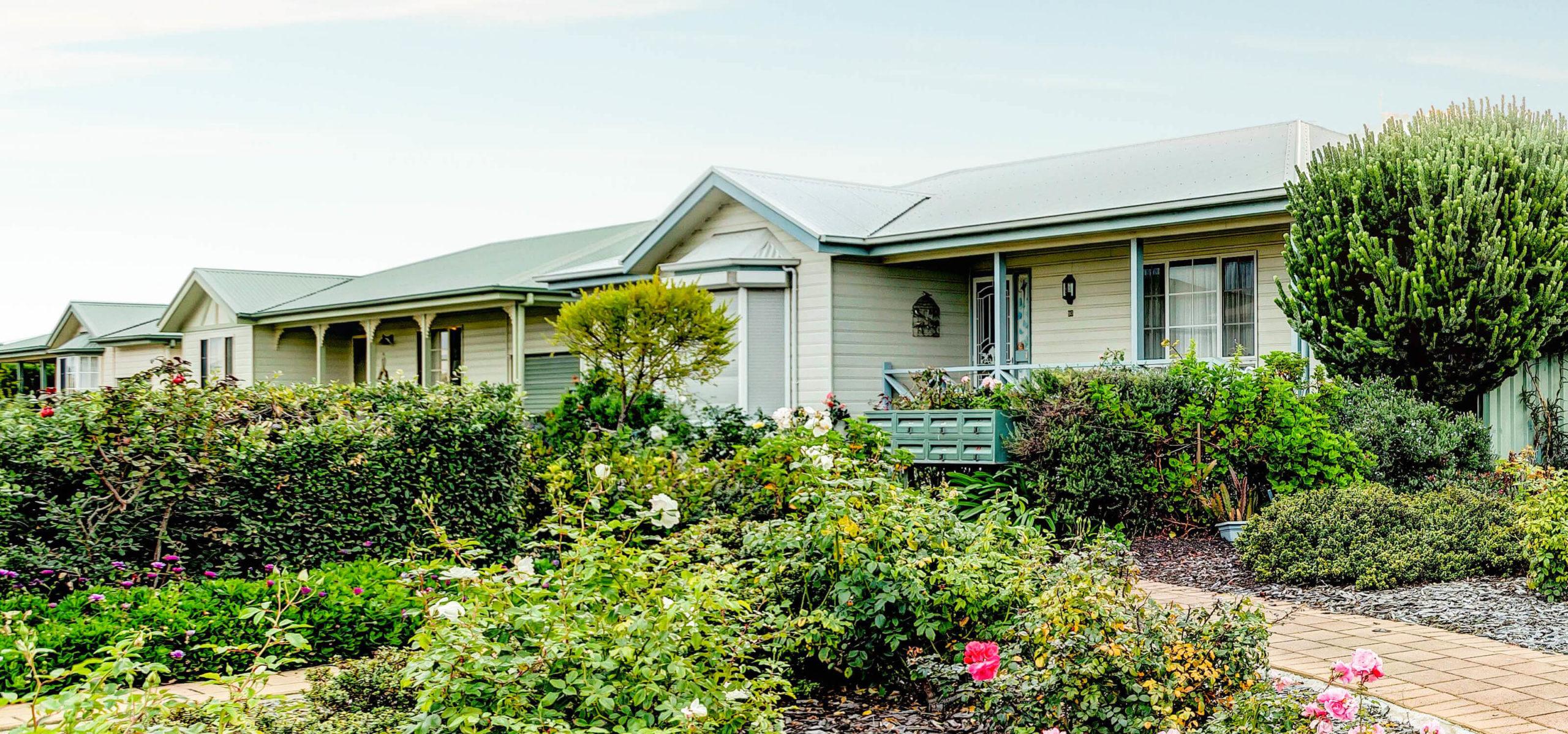 Single-story lifestyle village house with light exterior among lush green bushes and vibrant flowers, pathway leading to entrance in tranquil suburban environment.