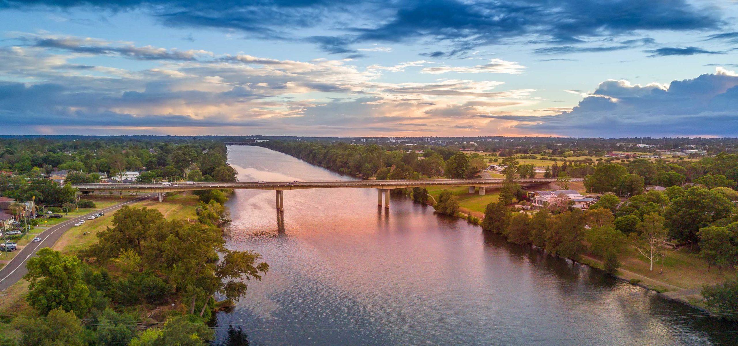Bridge spanning a wide river reflecting the sky at sunset, with roads, greenery, and potential lifestyle village houses lining the banks.